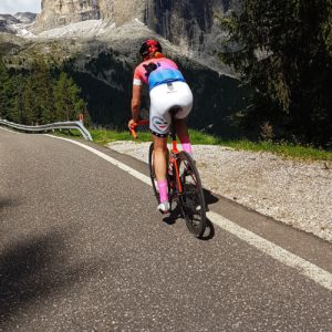 Woman wearing white bike shorts and a pink and blue jersey cycling on a red bike along the side of a road.