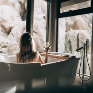 Woman doing self-care routine of sitting in a bathtub with a glass of wine while looking out windows.