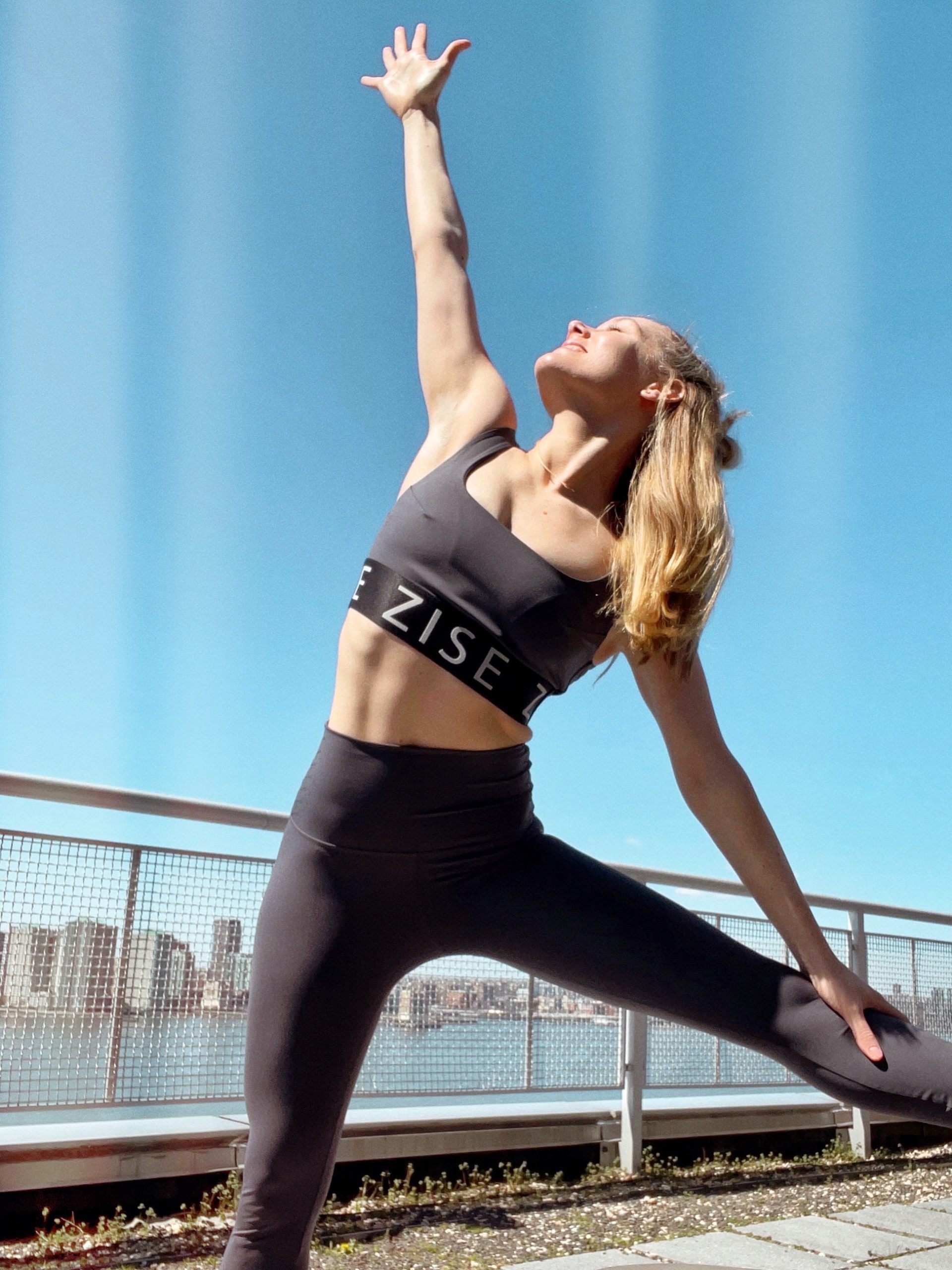 A woman wearing black ZISE workout clothes stretching her legs while reaching one arm to the sky