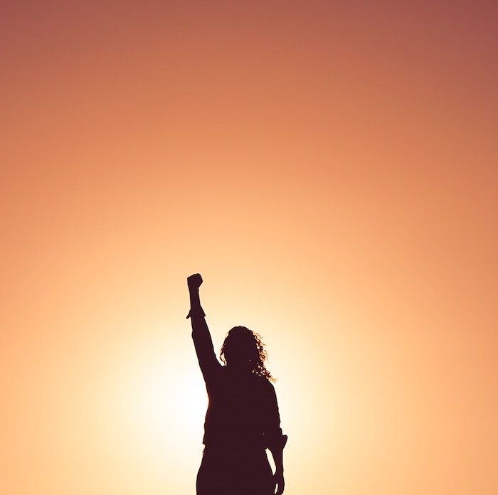 A silhouette of a woman standing in front of a sunset with her fist in the air.